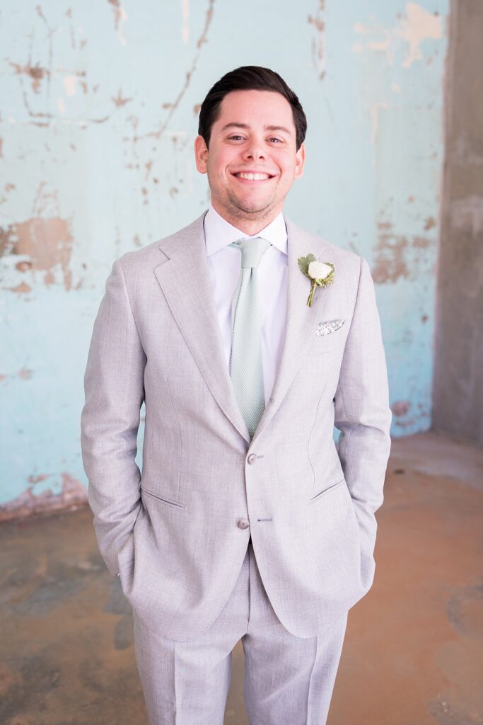 A dapper groom in a classic black suit and tie, posing against the rustic backdrop of Judson Mill in downtown Greenville, South Carolina.
