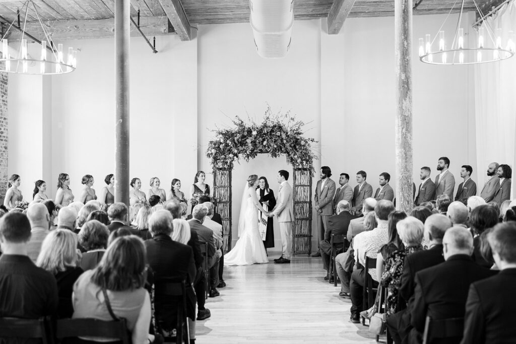 The bride and groom stand hand in hand, exchanging heartfelt vows in front of their loved ones at Judson Mill.