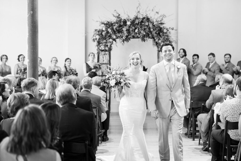 The bride's elegant white gown gracefully flows behind her as she walks arm-in-arm with her beaming groom up the aisle at Judson Mill wedding in downtown Greenville, South Carolina.